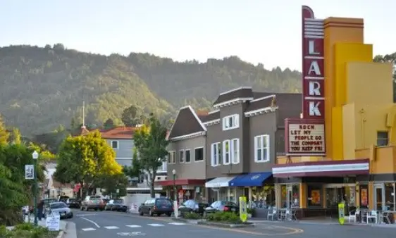 Street scene in a small town with a tall red LARK theater sign, colorful storefronts, cars, and green hills in the background.