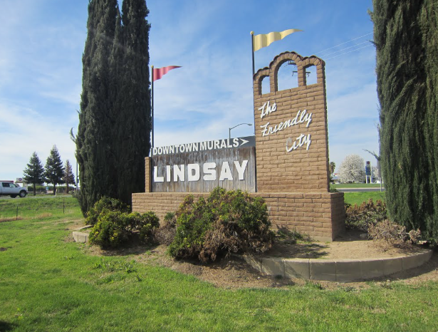 Welcome sign for Lindsay, 'The Friendly City,' with 'Downtown Murals' and large 'LINDSAY' lettering, set in brick monument with greenery nearby.