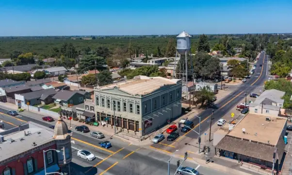 Aerial view of a small-town intersection; a green historic building sits on the corner with storefronts and a tall water tower nearby.