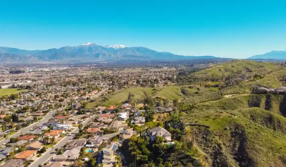 Aerial view of a suburban neighborhood with houses and streets at the base of green hills, mountains in the distance under a clear blue sky.